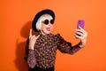 Photo of smiling cheerful happy modern grandmother take selfie on phone show rock n roll sign isolated on orange color