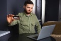 Photo of smiling man 30s in casual wear sitting alone in apartment and using laptop with credit card for shopping online Royalty Free Stock Photo