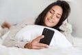 Photo of smiling woman 30s using mobile phone, while lying in bed with white linen in bright room Royalty Free Stock Photo