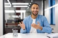 Photo of a smiling young hispanic man in a denim shirt sitting at a desk in a happy office and talking to the camera via Royalty Free Stock Photo