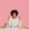 Photo of stupefied curly female designer wears white sweater and glasses, points with both index fingers at ceiling