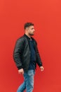 Photo of a stylish man with a beard in a leather jacket on a walk, walking on a background of a red wall, side view. Vertical Royalty Free Stock Photo