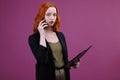 Photo of successful woman 20s holding clipboard with files in office and talking on mobile phone over pink background Royalty Free Stock Photo