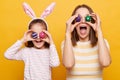 Photo of surprised cheerful positive mother and daughter in bunny ears covering eyes with Easter eggs, having fun together, family