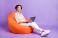 Photo of sweet charming young woman wear knitted shirt sitting beanbag typing modern gadget violet color