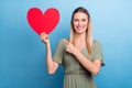 Photo of sweet shiny young woman dressed green clothes pointing finger large red heart card empty space smiling isolated