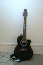 A dark colored acoustic guitar with shoulder strap resting upright against a white wall