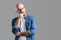 Portrait of thinking bald beard man in glasses who holds fist on chin isolated on gray background