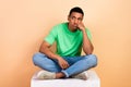 Photo of unhappy disapointed guy dressed green t-shirt sit on platform fist on cheek look empty space isolated on beige