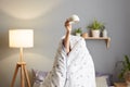 Photo of unknown unrecognizable woman sitting on bed wrapped in duvet hiding her head, putting her hand out of the blanket and Royalty Free Stock Photo