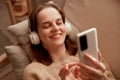 Photo of young adult Caucasian woman wearing headphones holding cell phone while lying on sofa in her apartment, using mobile Royalty Free Stock Photo