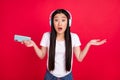 Photo of young asian girl unhappy shrug shoulders confused listen headphones hold cellphone isolated over red color Royalty Free Stock Photo