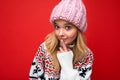 Photo of young beautiful blond lady with sincere emotions wearing pink knitted hat and winter pullover isolated over red Royalty Free Stock Photo