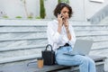 Beautiful thinking woman sitting outdoors using laptop computer talking by phone Royalty Free Stock Photo