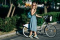 Photo of young blonde in long denim skirt standing next to bike on road next to green bushes in city Royalty Free Stock Photo