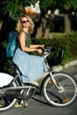 Photo of young blonde in sunglasses and long denim skirt standing next to bike near green bushes in city Royalty Free Stock Photo