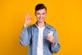 Photo of young cheerful man use mobile show fingers okey symbol promo isolated over yellow color background