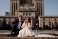 Photo of young couple in love, bride and groom in wedding dress are sitting on steps against backdrop of cityscape in sunglasses Royalty Free Stock Photo