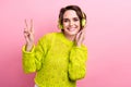 Photo of young girl in yellow jumper shows v sign promoting modern headphones control sound volume isolated on pink