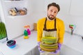 Photo of young handsome man hold pile clean plates tired wear apron housekeeper dirty duty kitchen indoors