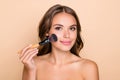 Photo of young happy beautiful girl applying highlighter blush on cheeks with makeup brush isolated on beige color