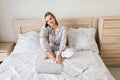 Photo of young joyful woman in pajama with laptop and smiling while sitting on bed in light room Royalty Free Stock Photo