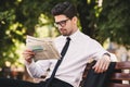 Photo of young man in businesslike suit sitting on bench in green park, and reading newspaper during sunny day Royalty Free Stock Photo