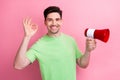 Photo of young smiling guy promoter holds megaphone shows okey sign to inform customers new products isolated on pink