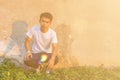 A young Thai boy is squating in front of dirty cement wall in the white shirt and jeans