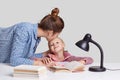 Photo of young woman cares of her child, kisses daughter in forehead, praises her to study well, explains material, read books and