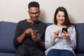 Photo of young woman in casual outfit sitting on couch in room with her African boyfriend and looking at theit smartphones`s on