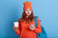 Photo of young woman feels good from received email, holds coffee to go, has backpack, carries rucksack, stands against blue wall Royalty Free Stock Photo