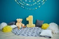 Photo zone with paper garlands, paper flowers, pillows, bear and number one on floor against blue background.