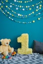 Photo zone with paper garlands, paper flowers, pillows, bear and number one on floor against blue background.