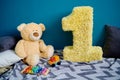 Photo zone with paper garlands, paper flowers, pillows, bear and number one on floor against blue background.