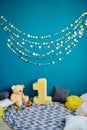 Photo zone with paper garlands, paper flowers, pillows, bear and number one on floor against blue background.