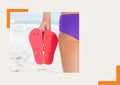 Photograph of mid section of woman holding flip flops at the beach against grey background