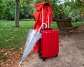 Photograph of a travel suitcase, an umbrella and a red gabardine in a park