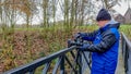 Photographer in blue jacket, hat and gloves on a bridge with his camera on a tripod to take a picture surrounded by trees Royalty Free Stock Photo