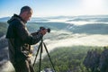 Photographer takes photos with camera on tripod on rocky mountain peak. Beautiful misty sunrise and valley view over Royalty Free Stock Photo