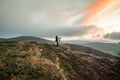 Photographer on top summit of mountain hill at dawn as sunrise illuminates clouds in sky beautiful orange, using a tripod. Royalty Free Stock Photo