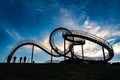 Photographers with tripods as silhouettes at the walkable roller coaster sculpture Tiger and Turtle against a dark blue sky with