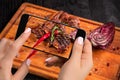 Photographing food concept - tourist takes picture of ready-to-eat beef steak dish on cutting wooden board on smartphone