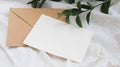 Photostock wedding styled composition. Feminine envelope mockup scene with ruscus leaves, silk ribbon, blank greeting card, on cre