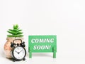 Phrase Coming Soon Written On Green Paper Card With Alarm Clock And House Plant.