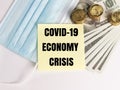 Phrase COVID-19 ECONOMY CRISIS on note book with dollar bank notes, coins and face mask.