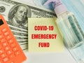 Phrase COVID-19 EMERGENCY FUND on sticky note with dollar bank notes, calculator, hand sanitizer and face mask.