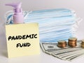 Phrase PANDEMIC FUND on sticky note with dollar bank notes ,coins,hand sanitizer and a bunch of face mask.