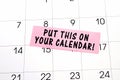 The phrase Put This on Your Calendar written on a pink sticky note posted on a calendar or planner page. Closeup of a personal