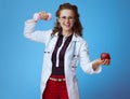 Physician woman with dumbbell and apple showing biceps on blue